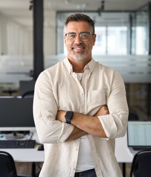 Handsome hispanic senior business man with crossed arms smiling at camera. Indian or latin confident mature good looking middle age leader male businessman on blur office background with copy space.